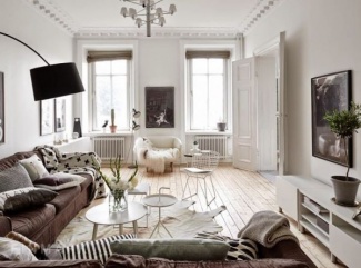 Ambiente total branco, com quadros escuros na parede, sofá marrom com muitas almofadas e mantas. Piso de madeira clara.